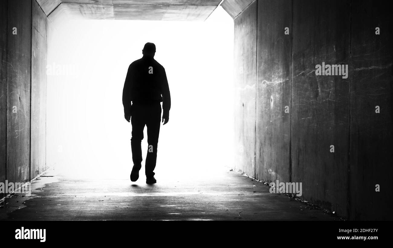 silhouette of man, shoulders slumped walking out of a dark tunnel towards a bright light. Stock Photo