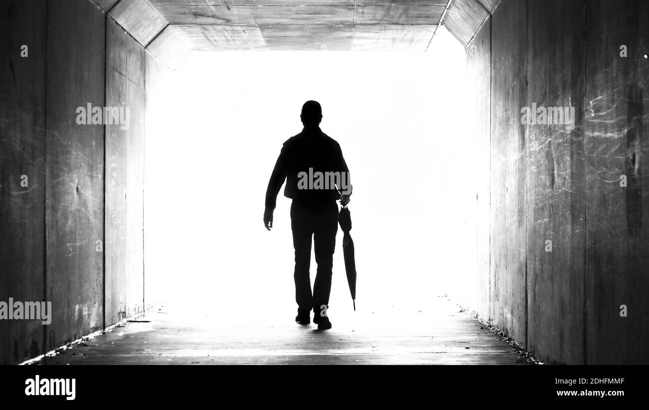 A silhouette of a man holding a folded up umbrella leaving a dark tunnel walking towards the light. Weather, climate, leaving and departure theme Stock Photo