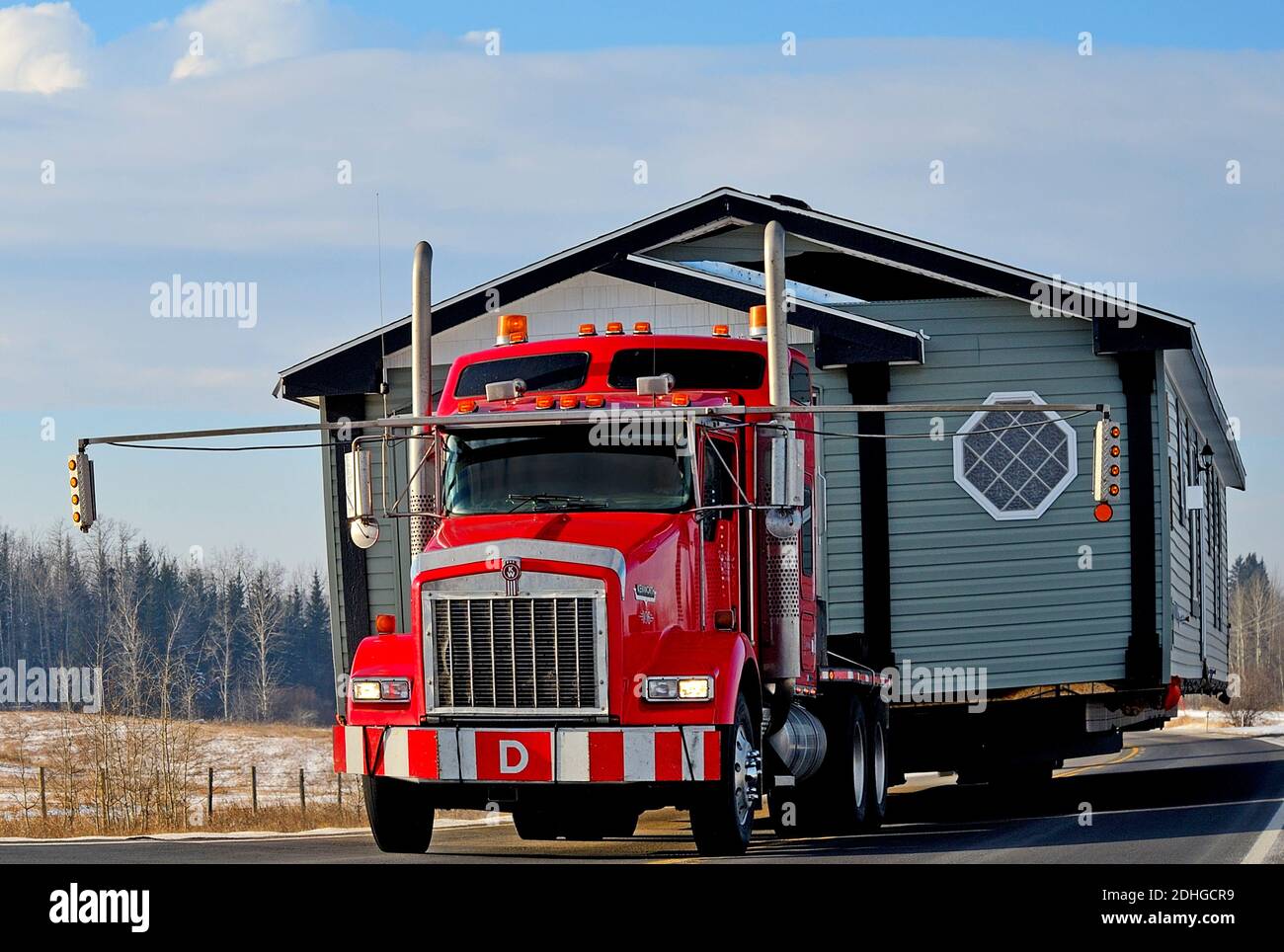 A close up image of a red semi truck moving a house along a two lane highway in rural Alberta Canada Stock Photo