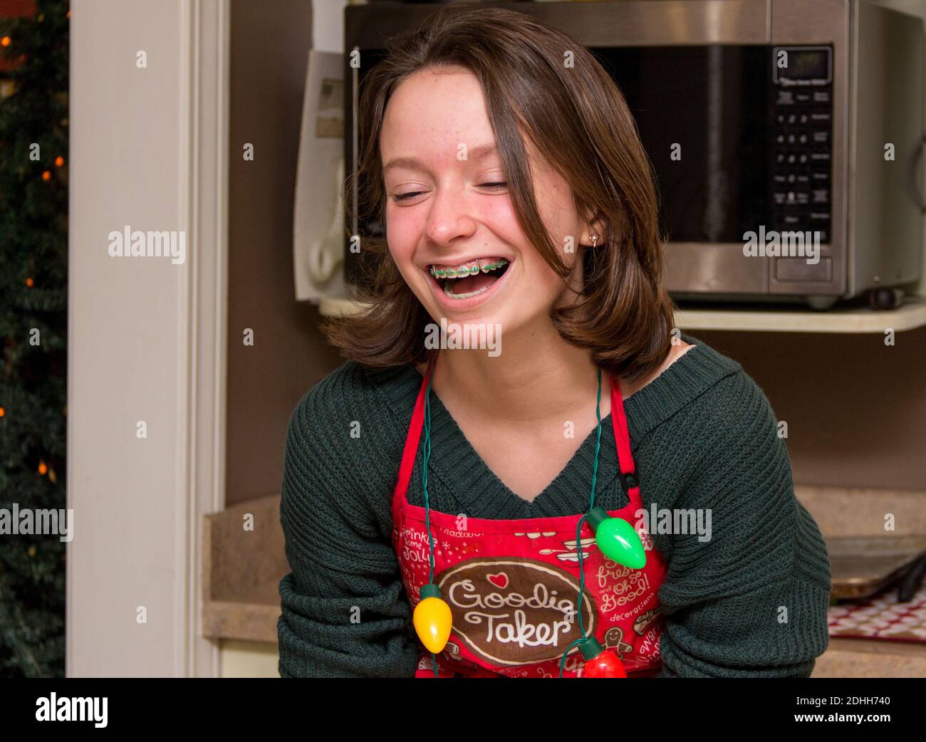 Young girl laughing in green sweater with christmas apron on in kitchen of house. Girl is wearing a necklace of fake christmas lights around her neck. Stock Photo
