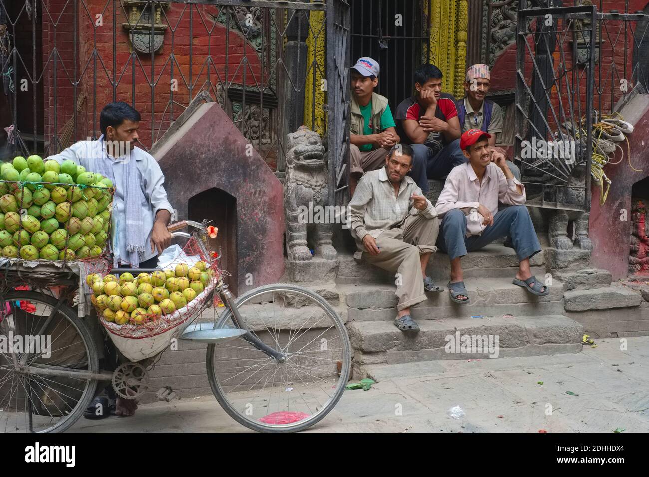 A mobile fruit vendor with his bicycle, in front of a small temple, with a bunch of men idly idling on its steps - a street scene in Kathmandu, Nepal Stock Photo
