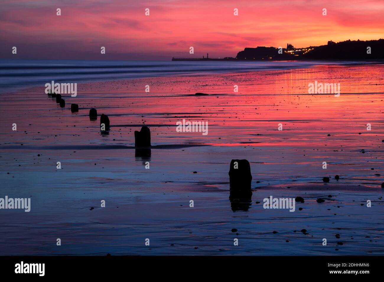 Daybreak view looking towards Whitby showing pier, town and abbey in silhouette against a colourful sky reflected on wet sand.  Viewed from Sandsend. Stock Photo