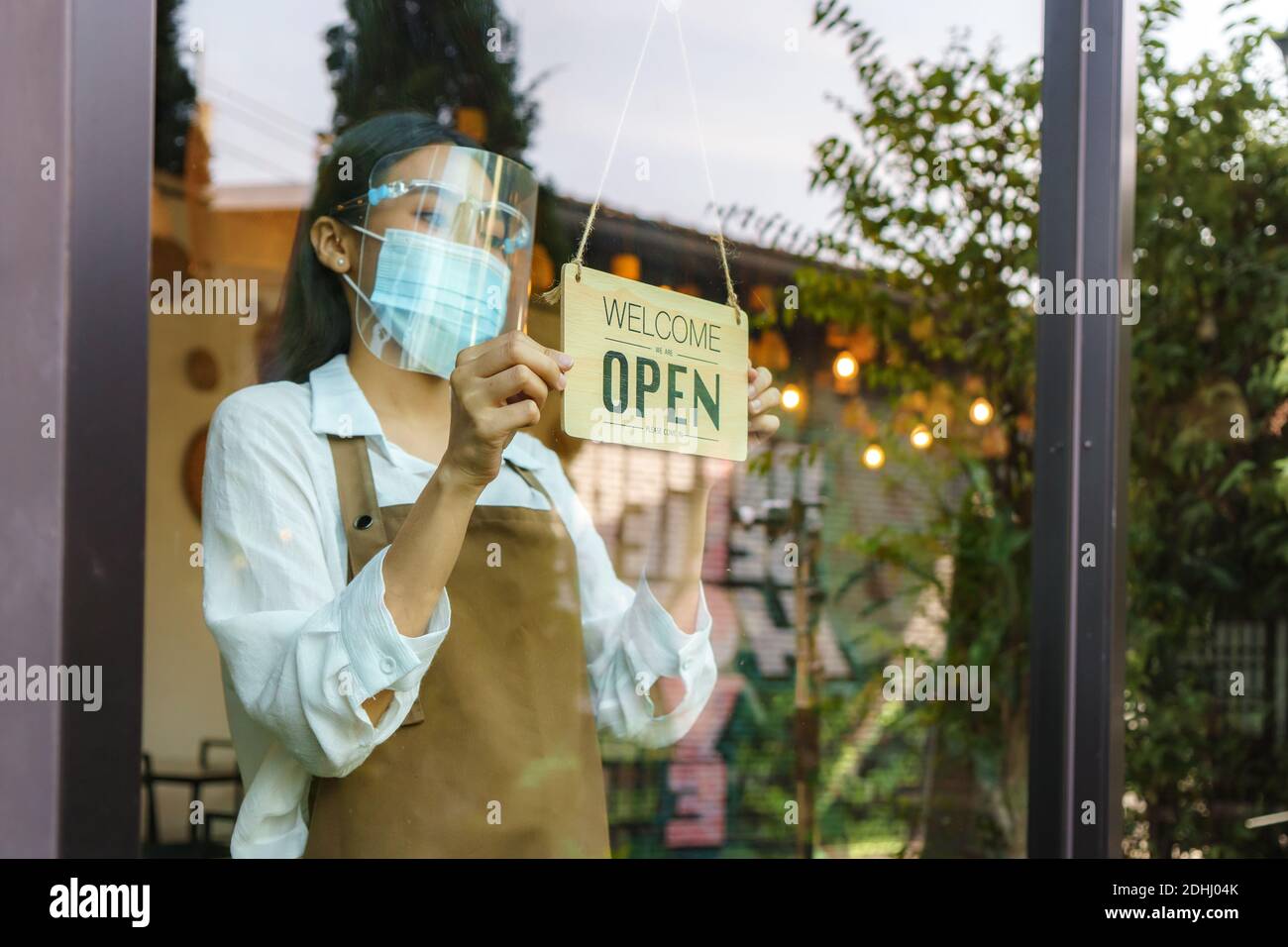 Asian business owner woman wear protective face mask and face shield hanging open sign at her restaurant / cafe, open again after lockdown for new nor Stock Photo