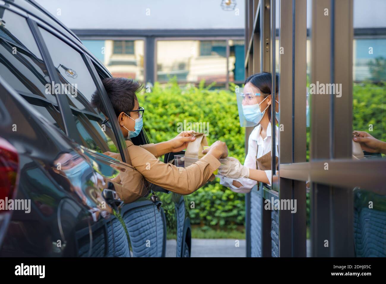 Asian man in protective mask taking food bag and coffee with woman waitress wearing face mask and face shield at drive thru during coronavirus outbrea Stock Photo