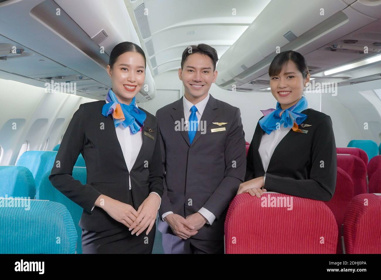 Portrait of three man and woman in blue suit flight attendant/air hostess in economy class cabin smiling to welcome passenger at airplane. Stock Photo