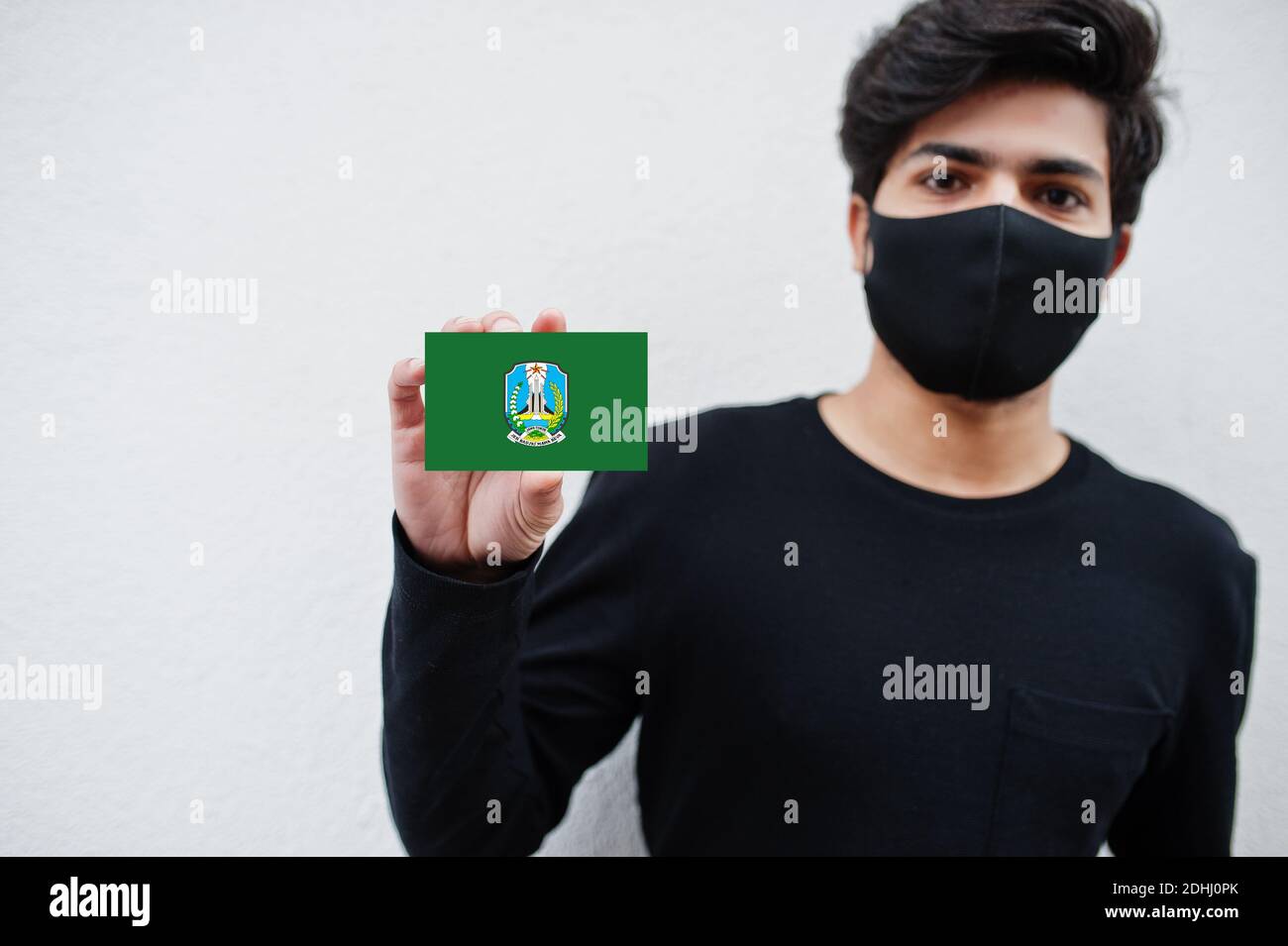 Indonesian man wear all black with face mask hold East Java flag in hand isolated on white background. Provinces of Indonesia coronavirus concept. Stock Photo
