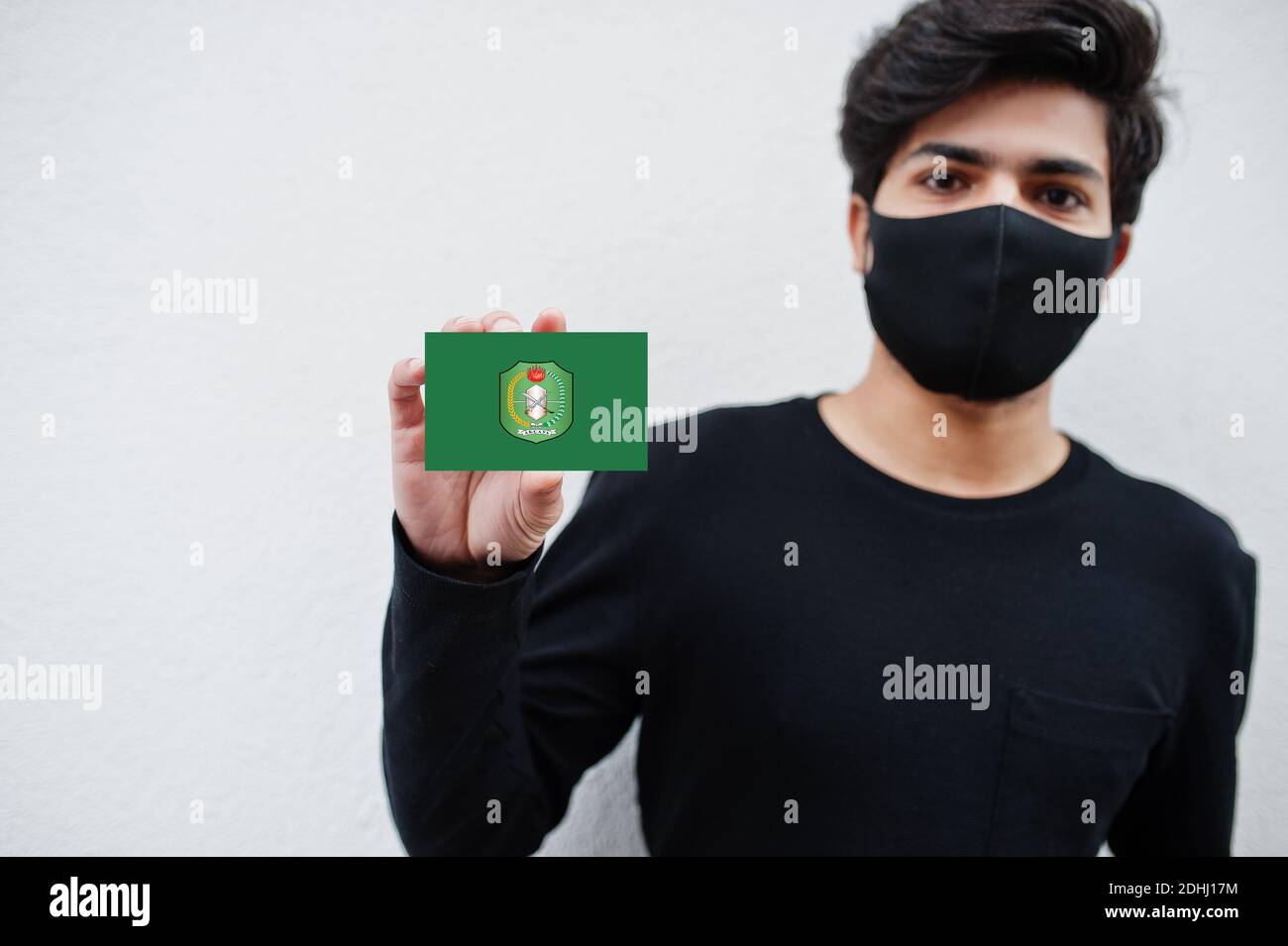 Indonesian man wear all black with face mask hold West Kalimantan flag in hand isolated on white background. Provinces of Indonesia coronavirus concep Stock Photo