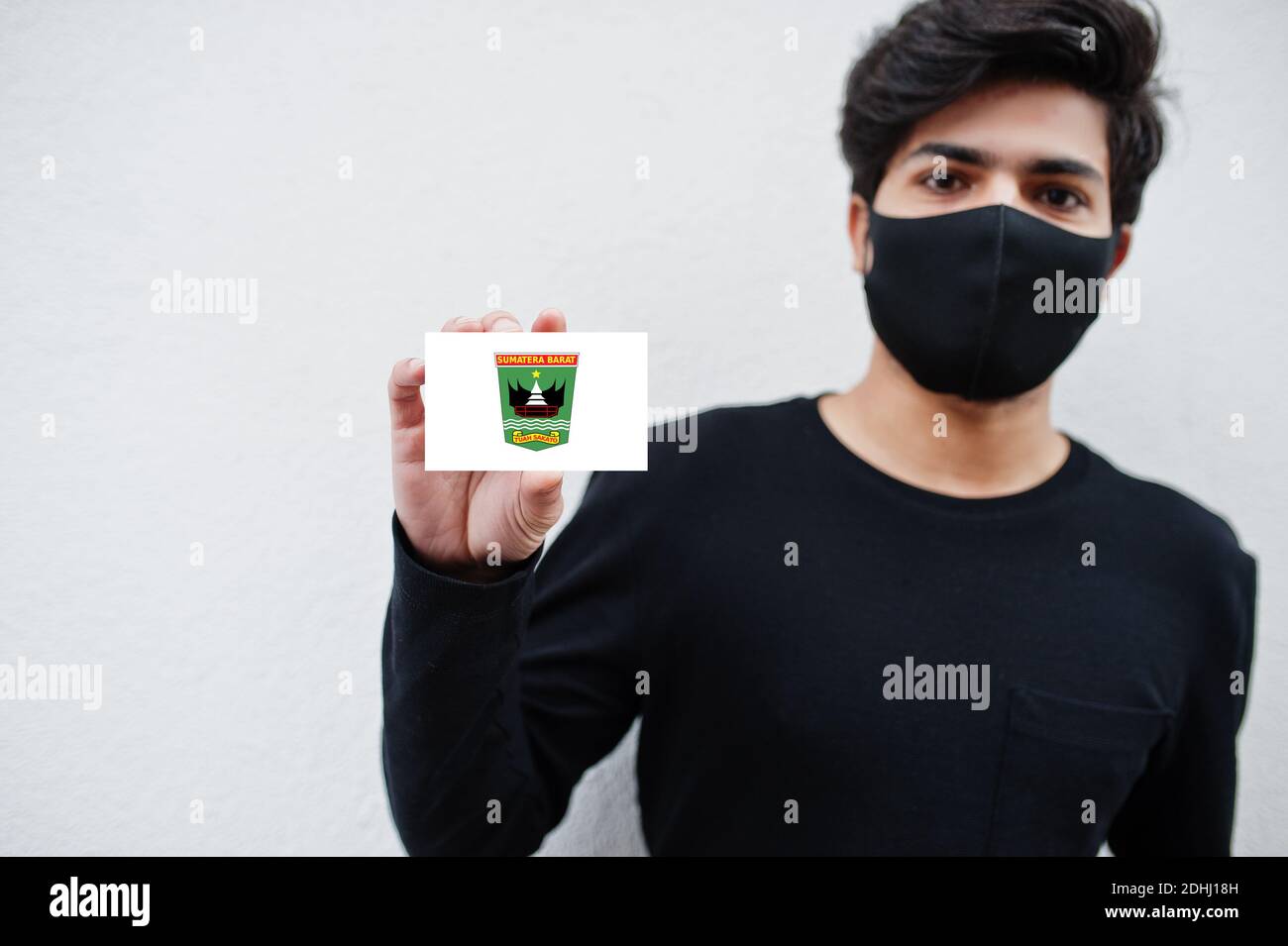 Indonesian man wear all black with face mask hold West Sumatra flag in hand isolated on white background. Provinces of Indonesia coronavirus concept. Stock Photo