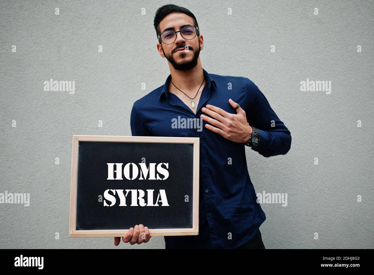 Arab man wear blue shirt and eyeglasses hold board with Homs Syria inscription. Largest cities in islamic world concept. Stock Photo