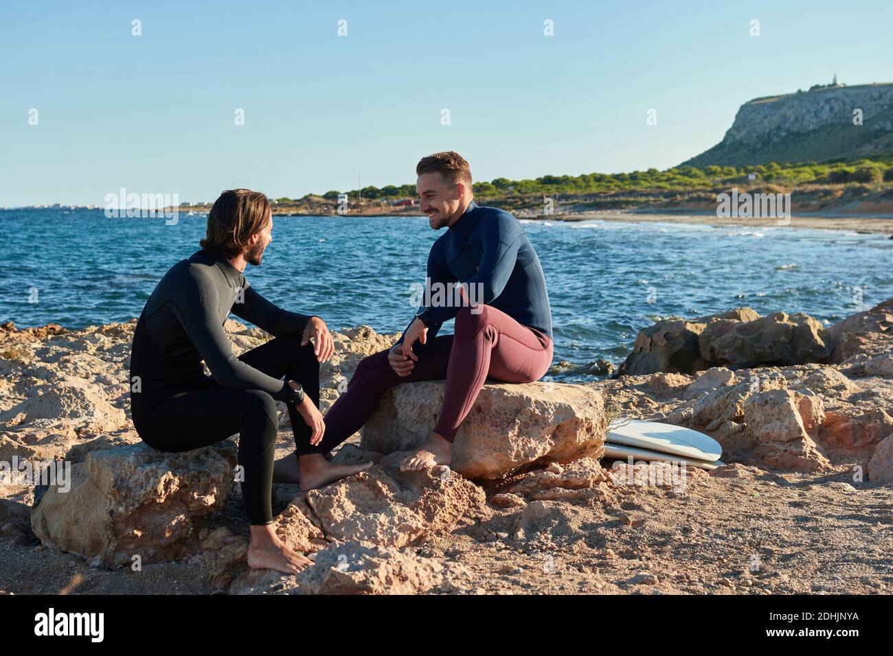 Cheerful male surfers in wetsuits sitting on rocks with surfboards in