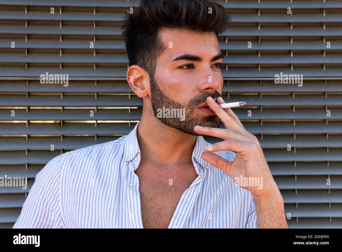 Thoughtful handsome male leaning on building and smoking cigarette in city while looking away in contemplation Stock Photo