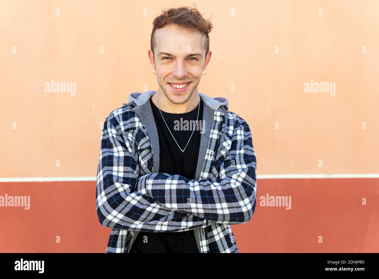 Positive confident young hipster guy in casual checkered shirt looking at camera and smiling while standing against orange wall Stock Photo