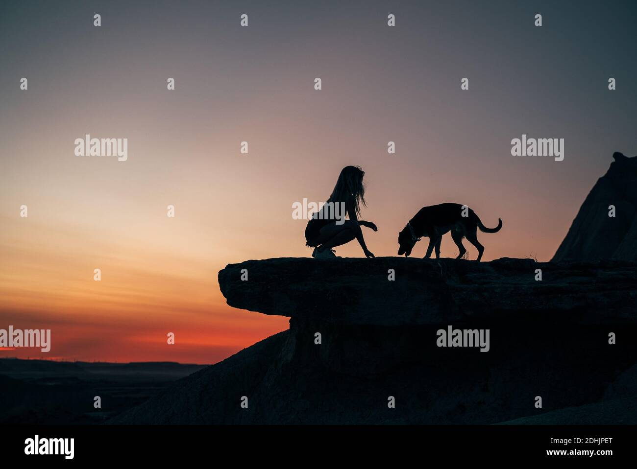 Side view of silhouette of unrecognizable female sitting on hill with dog on background of sundown in Bardenas Reales Stock Photo