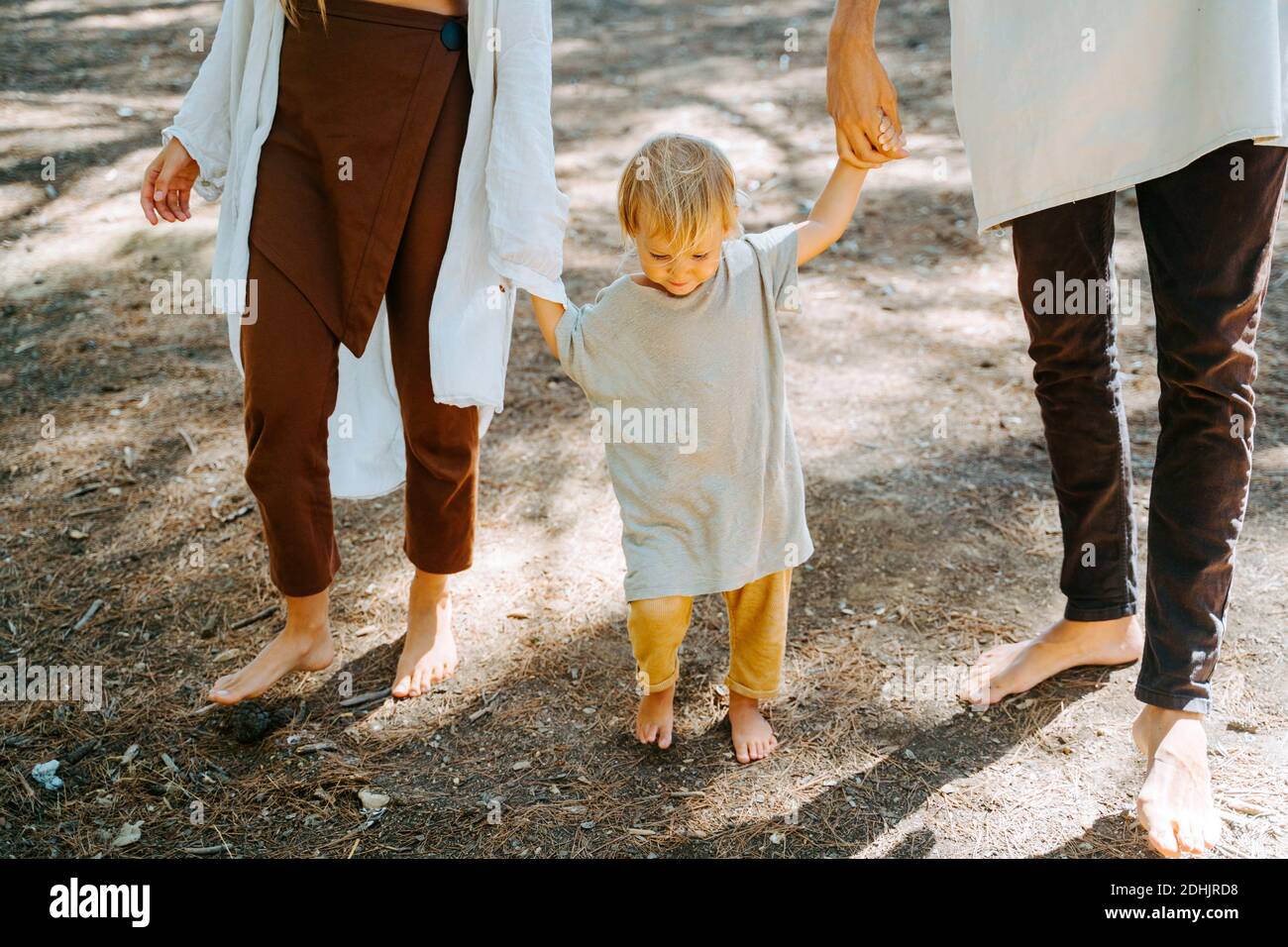 Cropped unrecognizable multiethnic hippie parents and cute toddler walking barefoot in woods on sunny day and enjoying unity with nature Stock Photo