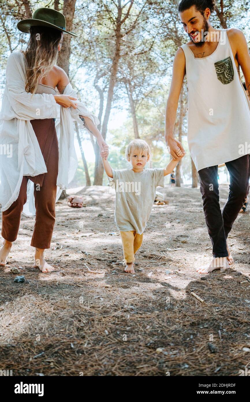 Family of multiethnic hippie parents and cute toddler walking barefoot in woods on sunny day and enjoying unity with nature Stock Photo