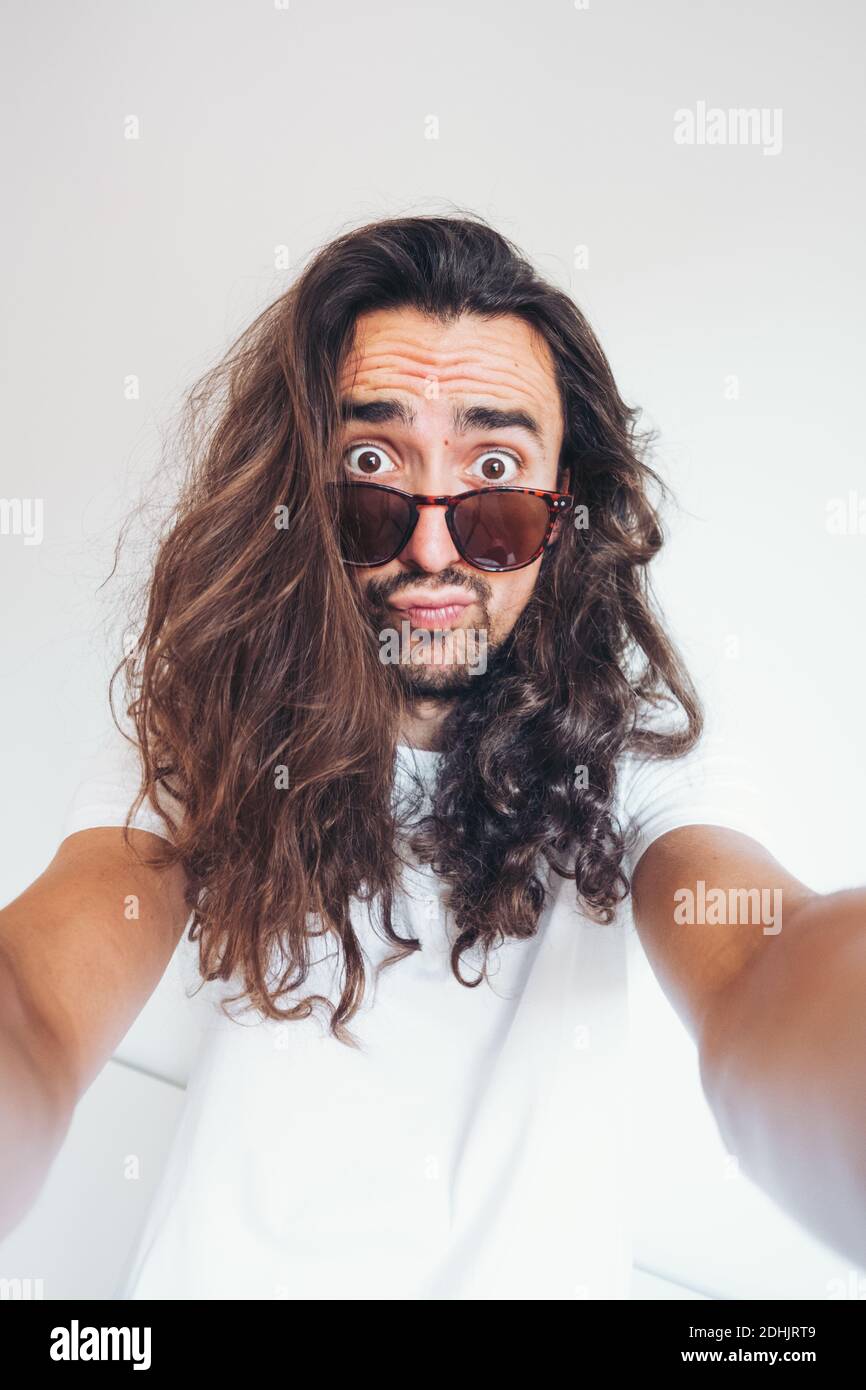 Young hipster long haired bearded male in sunglasses with funny amazed face expression looking at camera while taking selfie Stock Photo