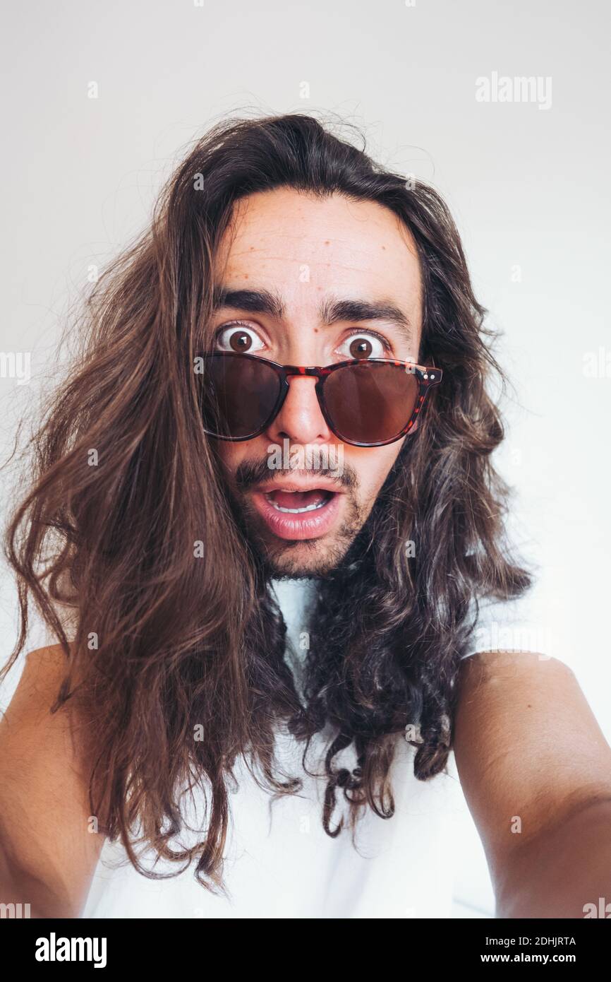 Young hipster long haired bearded male in sunglasses with funny amazed face expression looking at camera while taking selfie Stock Photo
