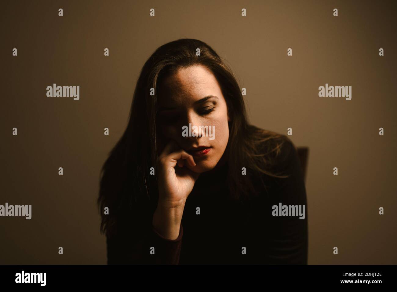 Unhappy pensive young female in black wear leaning on hand and thinking about personal problems while sitting against brown wall Stock Photo