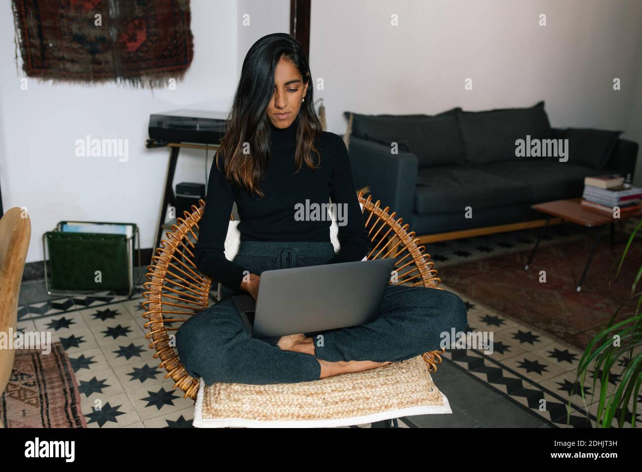 Serious Indian female freelancer sitting with crossed legs in wicker chair and typing on netbook while working online from home Stock Photo