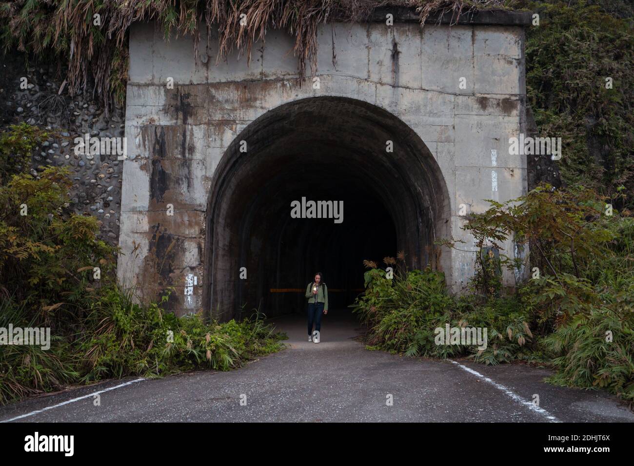 Distant unrecognizable traveler walking out of arched weathered stone tunnel on narrow asphalt road with green bushes on roadsides Stock Photo