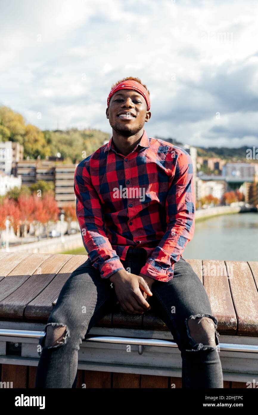 Confident smiling young African American guy in checkered shirt and ripped jeans sitting on fence near urban embankment and looking at camera Stock Photo Alamy