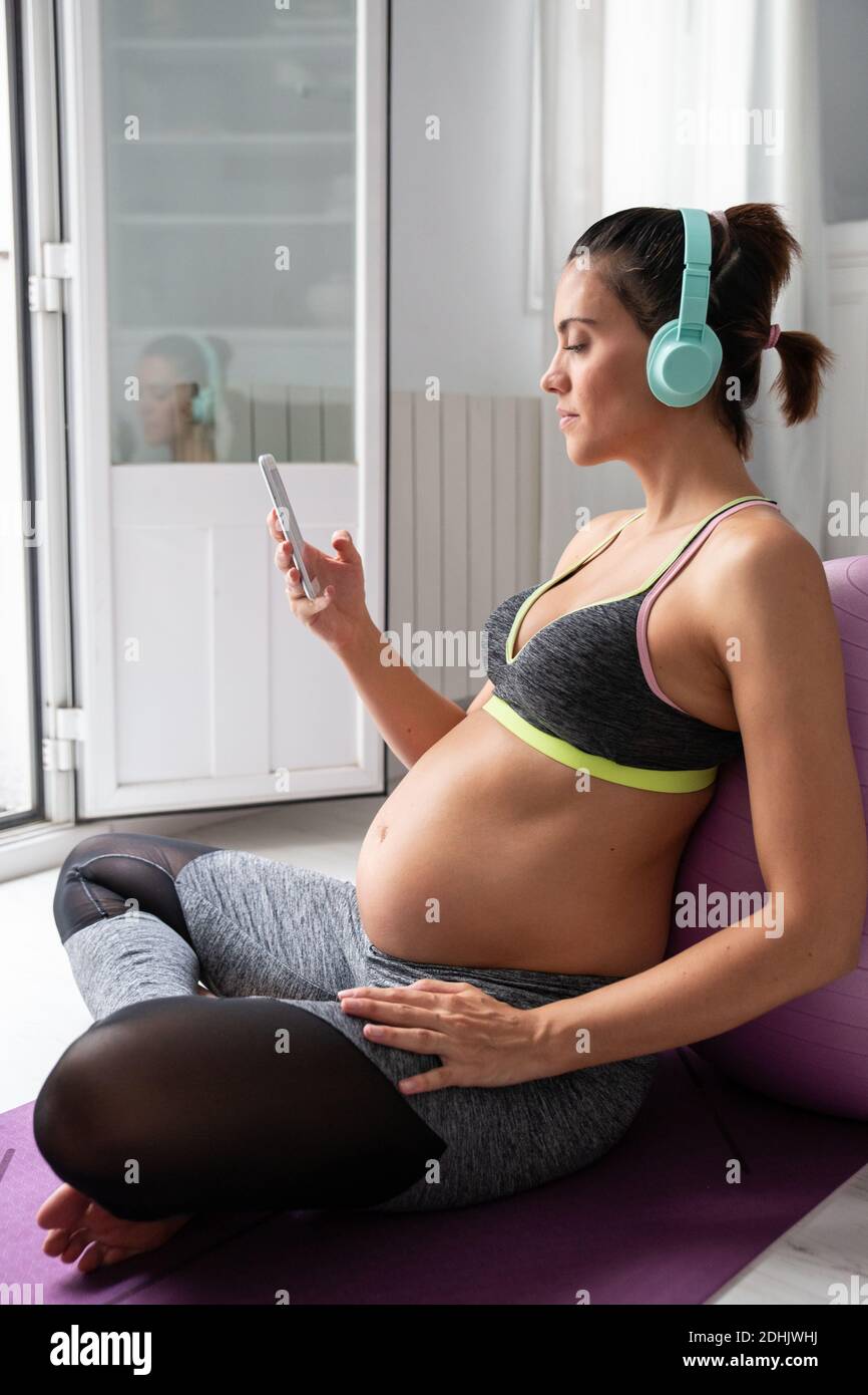 Side view of pregnant female sitting on mat using smartphone while listening to music in headphones after practicing yoga Stock Photo