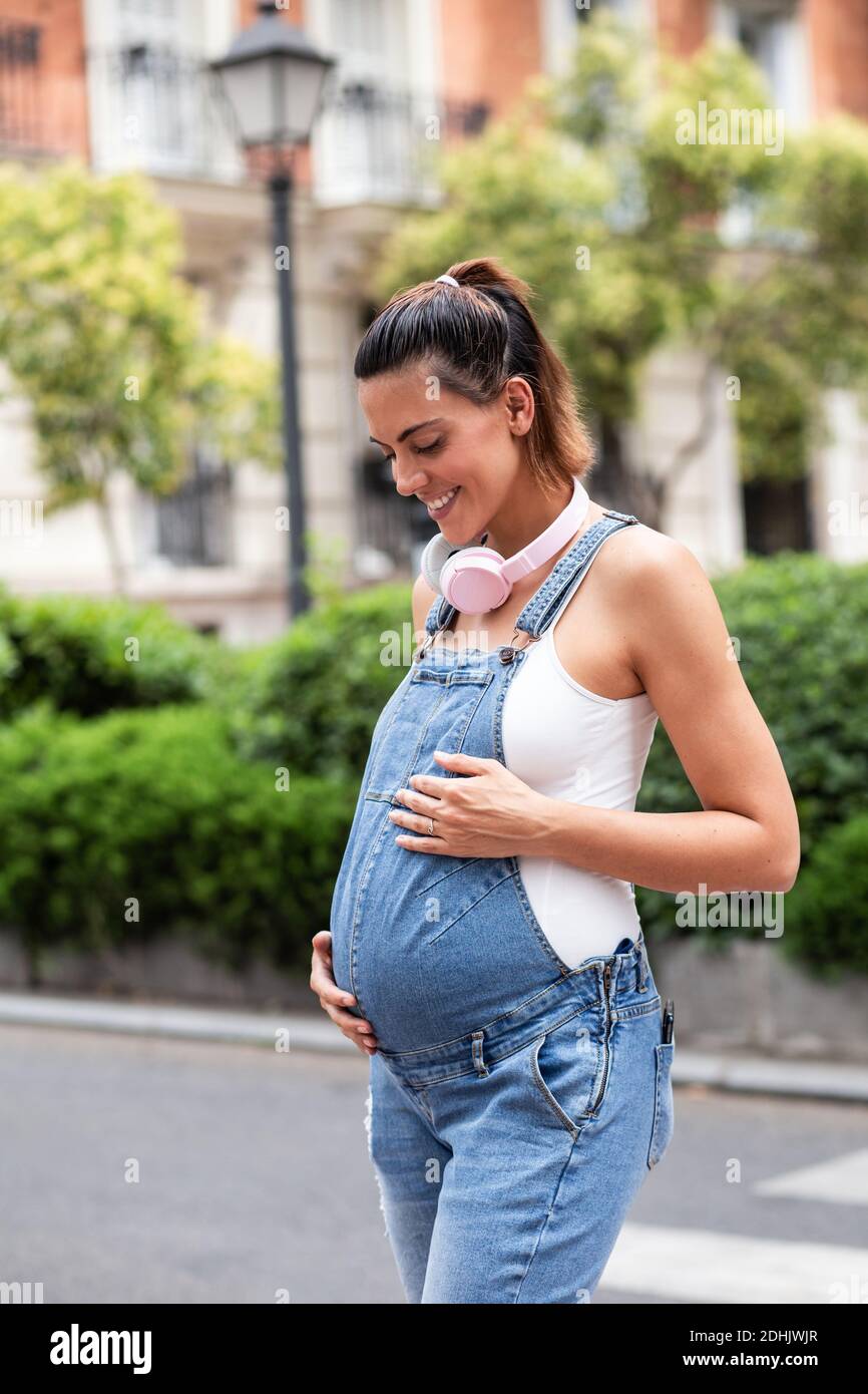 Side view of dreamy pregnant female in overalls standing on street
