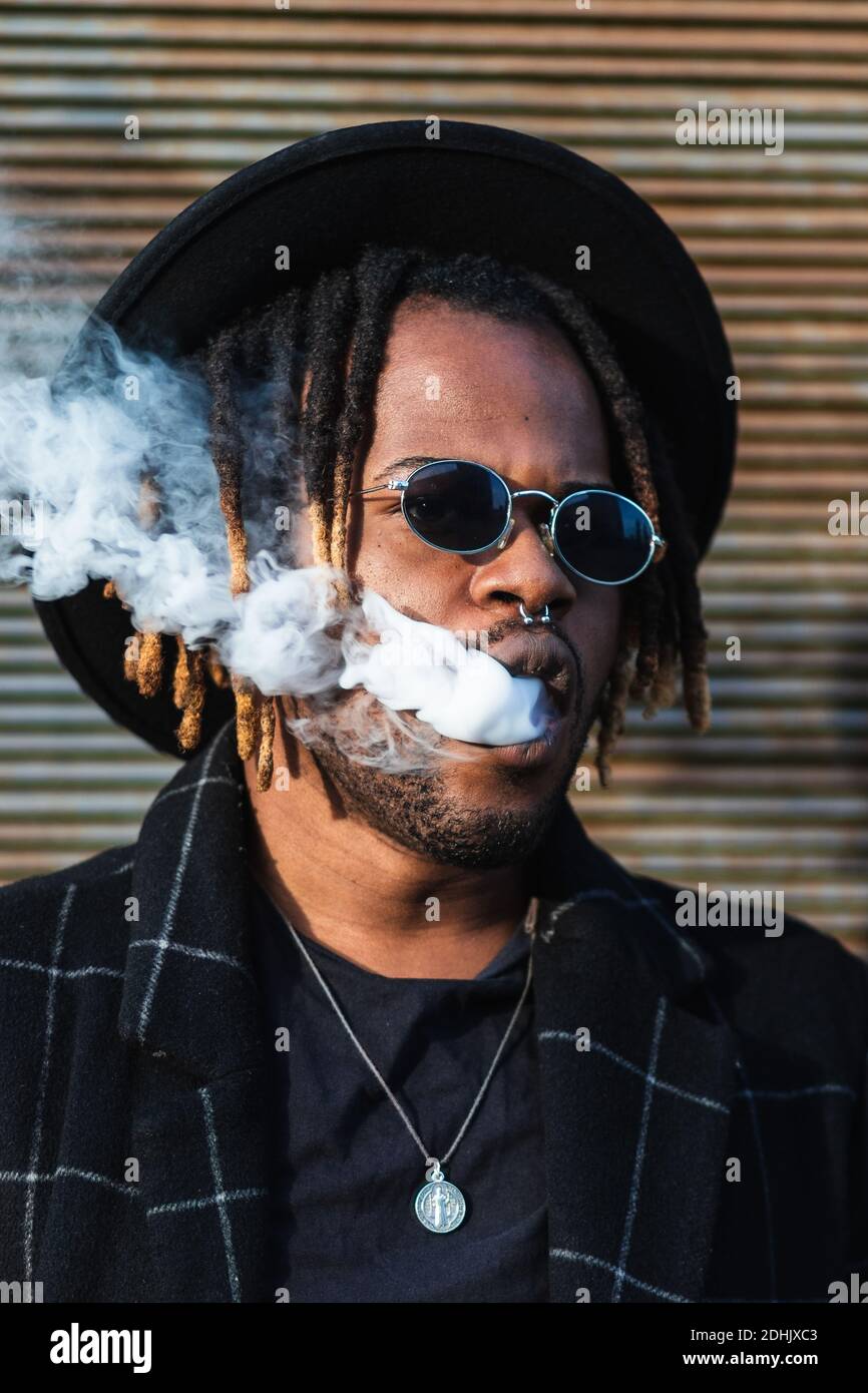 Modern African American man with dreadlocks wearing hat with sunglasses and exhaling smoke on street Stock Photo