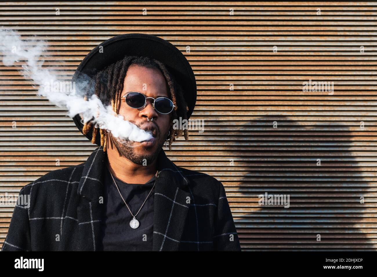 Modern African American man with dreadlocks wearing hat with sunglasses and exhaling smoke on street Stock Photo