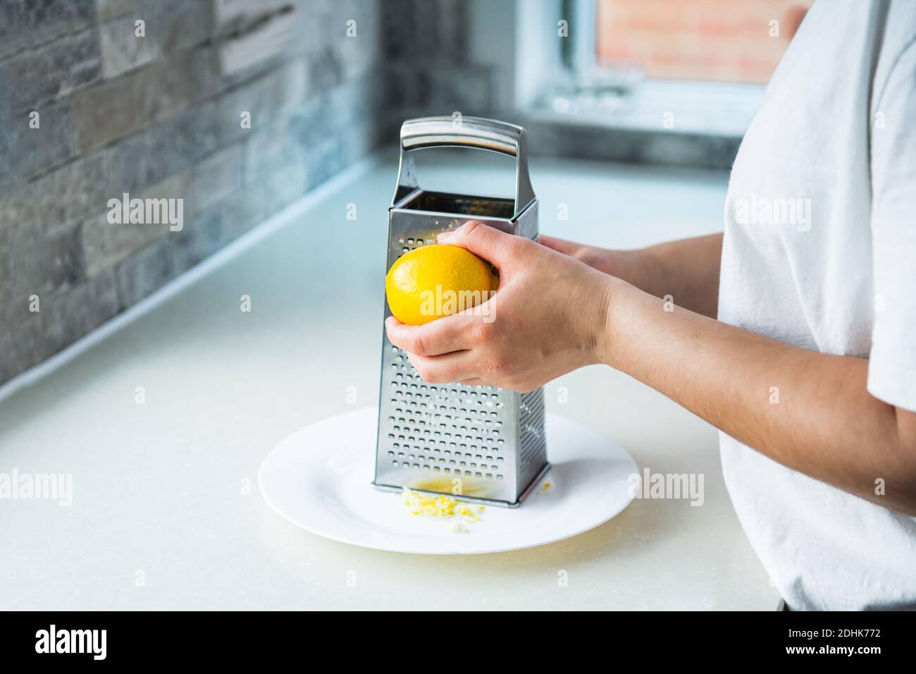 Female hands grating fresh whole lemon on metal hand grater, making zest on her kitchen table. Yellow and gray trendy colors of the year 2021 Stock Photo