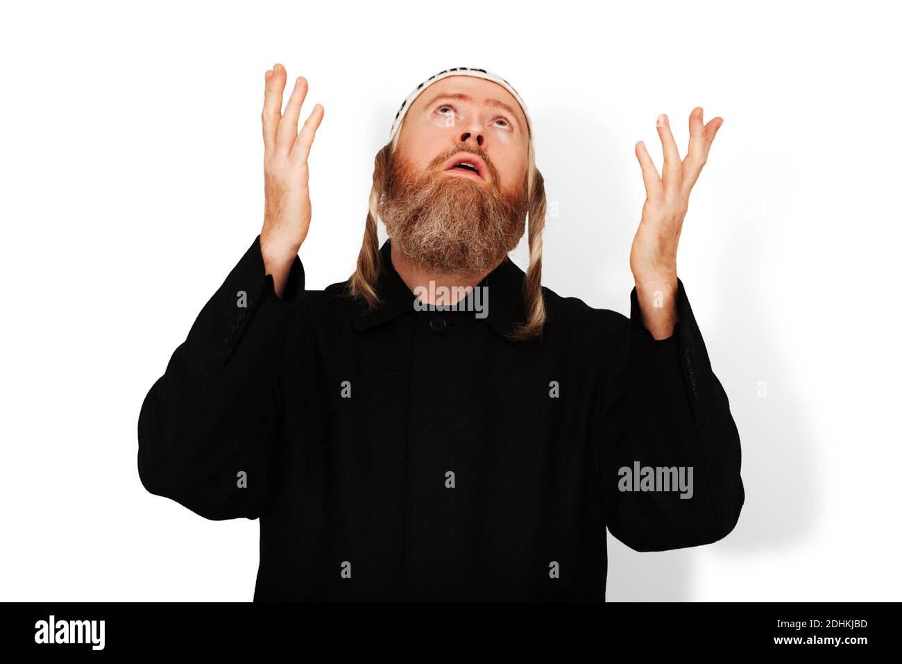 Praying bearded Jewish man with sidelocks in white kippah raising his hands to heaven. Studio portrait of hasid wearing black coat isolated on white Stock Photo
