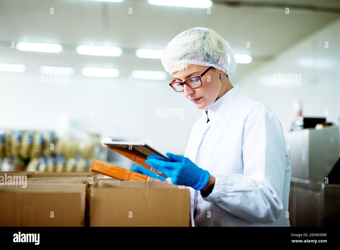 Young beautiful dedicated female worker in sterile clothes is carefully inspecting info on tablet and comparing it to the inventory inside of factory Stock Photo