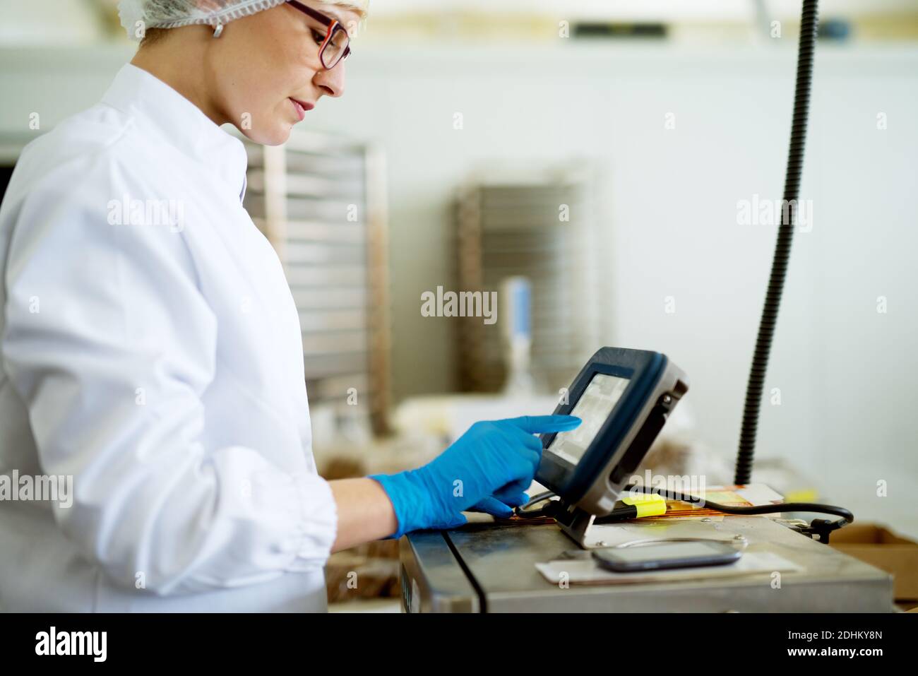 Young focused female worker in sterile cloths using a complex machinery in factory production line. Stock Photo