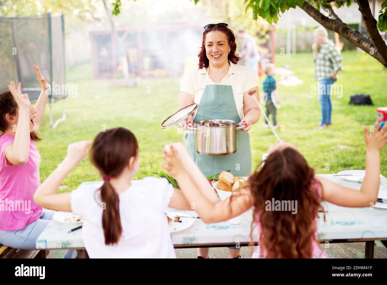 Playful little girls are sitting at the picnic table and are cheering for their mother who is bringing pot filled freshly grilled meat. Stock Photo