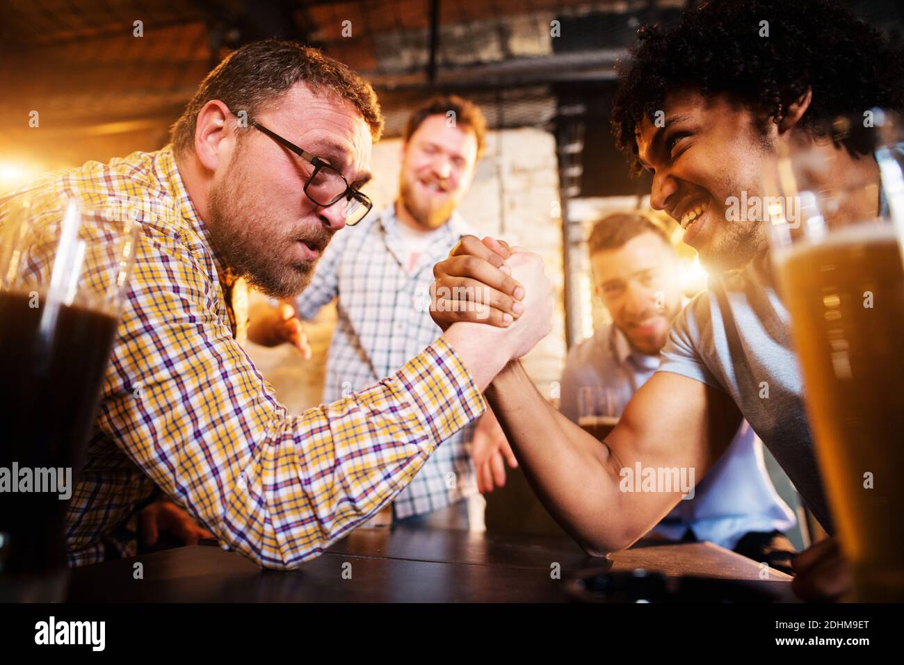 Exhausted mixed-race male friends having arm wrestling challenge at the local bar while other supporting. Stock Photo