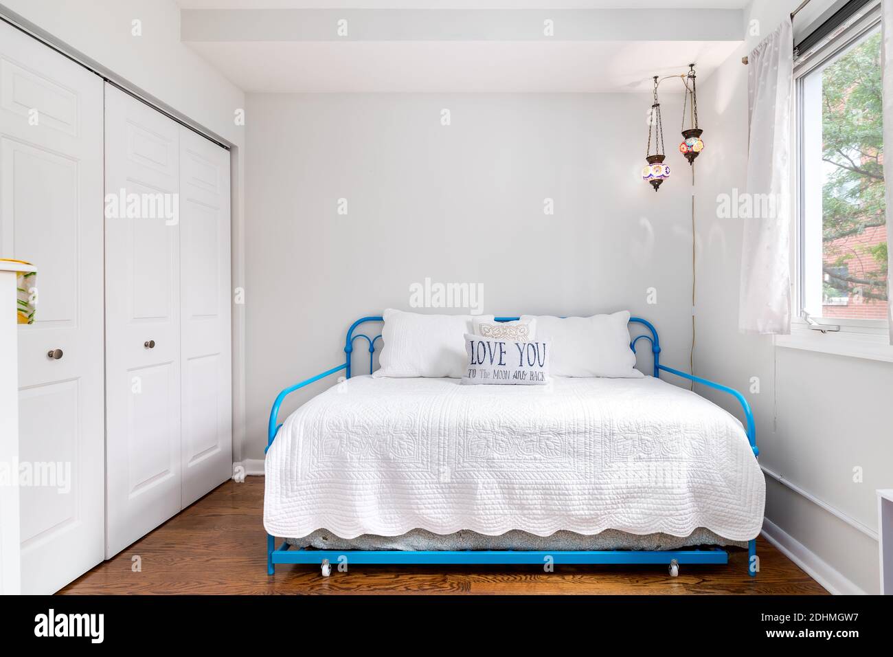 A small bedroom with a twin side bed, baby blue bed rails, a pillow that says, 'Love you to the moon and back,' and stained glass lights hanging. Stock Photo