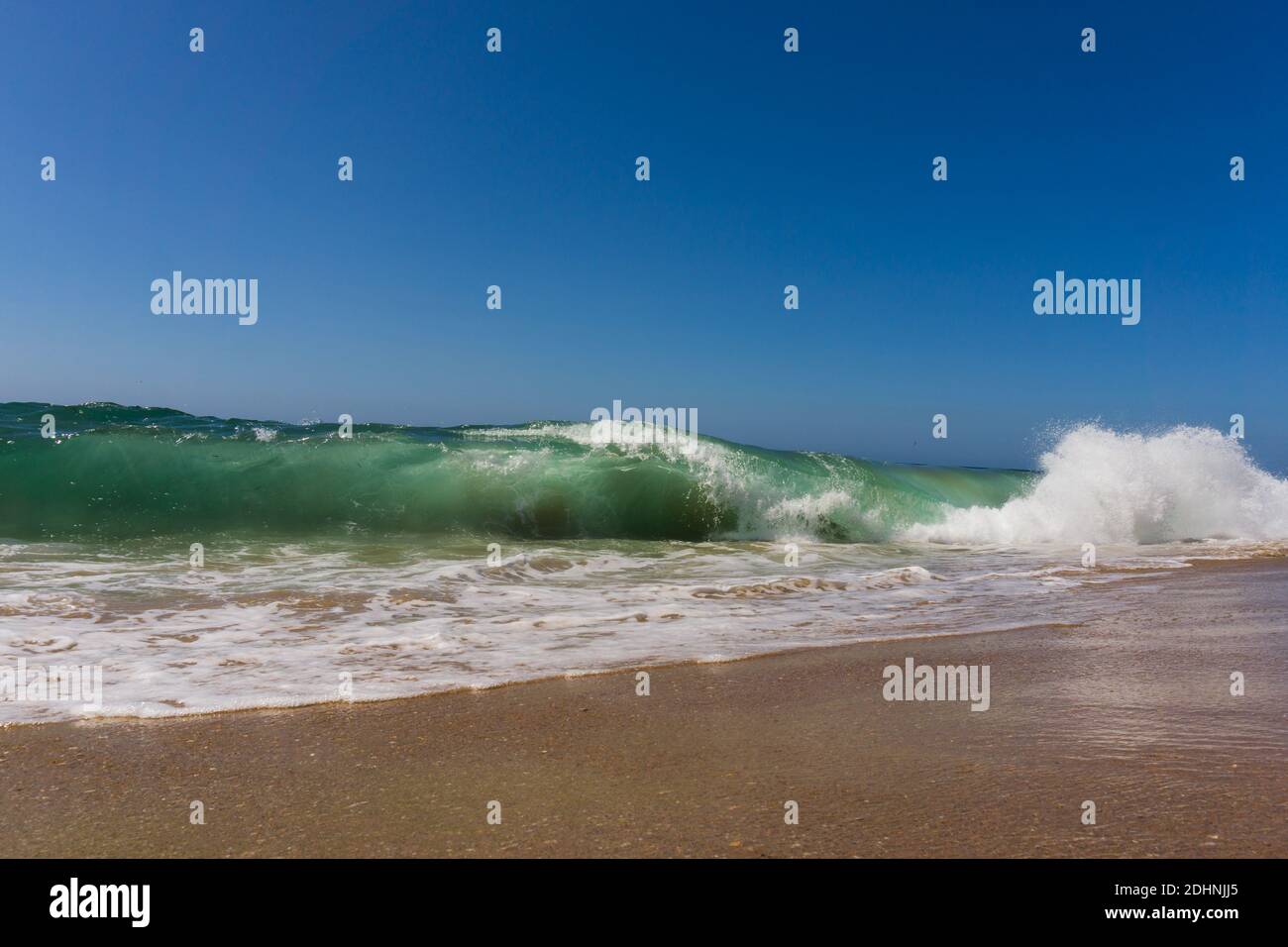 A wave curling over and crashing onto sandy beach under a blue sunny sky Stock Photo