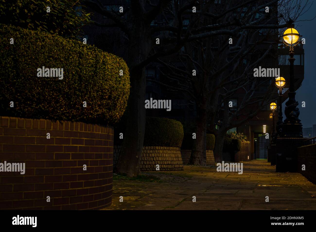 Nighttime on a pathway along red brick wall, hedges and street lamps leaving yellow glow Stock Photo