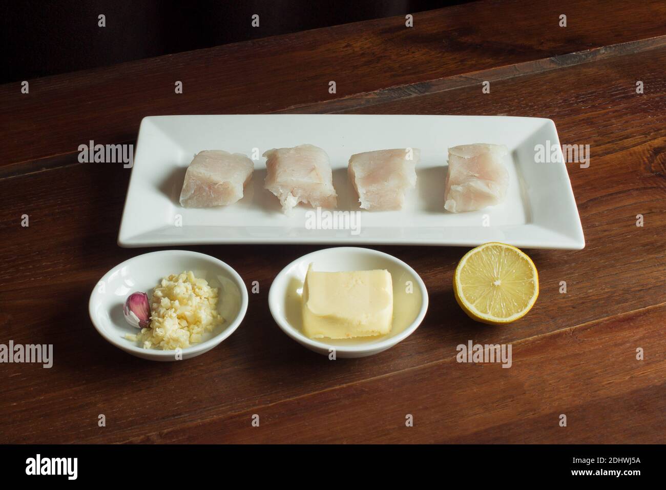 Food plating and presentation of raw food on an old wooden table. Hake, butter, garlic, and half a lemon. Stock Photo
