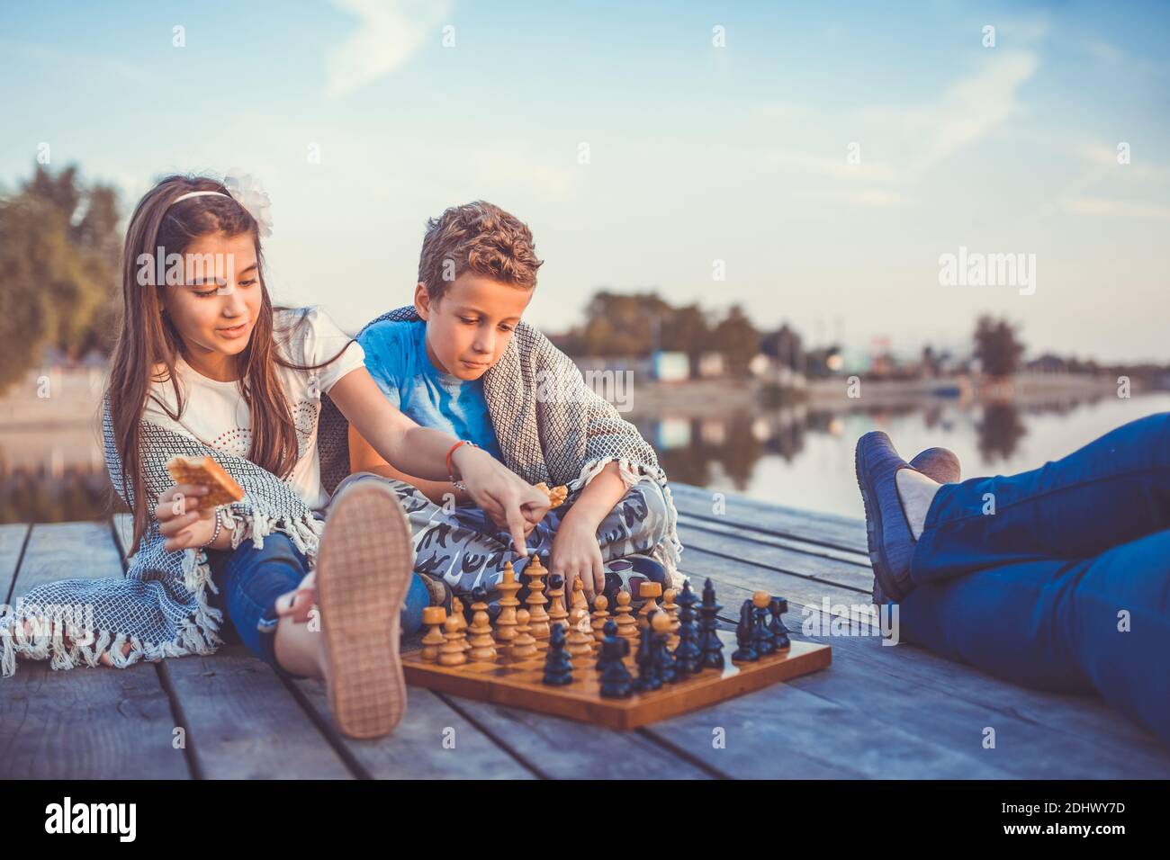 Two young cute little friends, boy and girl having fun while playing chess sitting by the lake in the evening. Kids are playing. Friendship. Selective Stock Photo