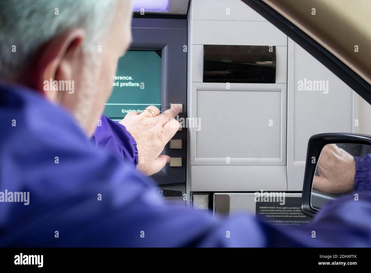 Grey-haired man at drive-up ATM machine leaning out car window and pushing button - Instructions in English and Spanish - Selective focus. Stock Photo