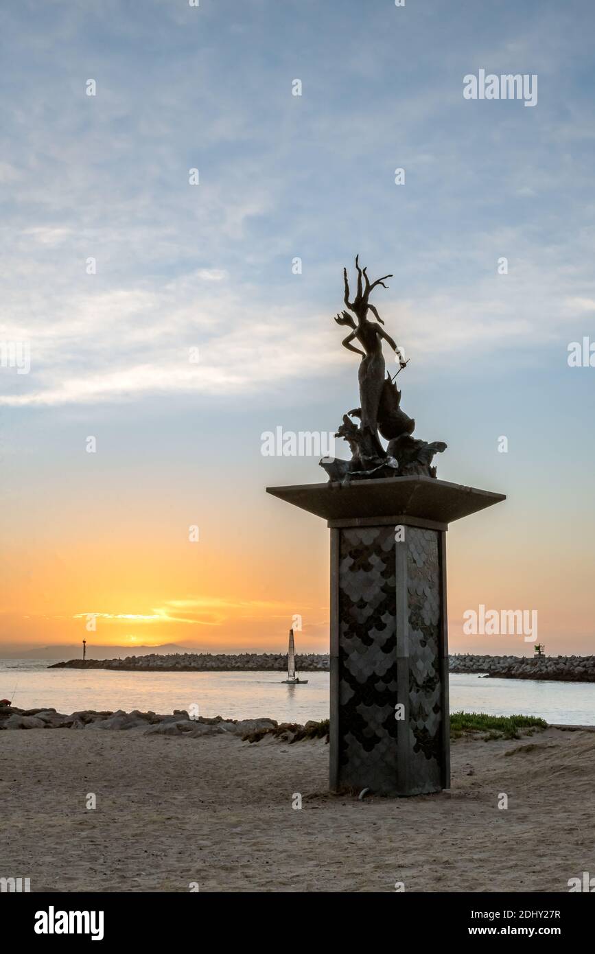 Ventura harbor mouth entrance has mermaid statue ushering the lone sailboard into the safety of harbor as sun sets behind island horizon of Pacific Oc Stock Photo