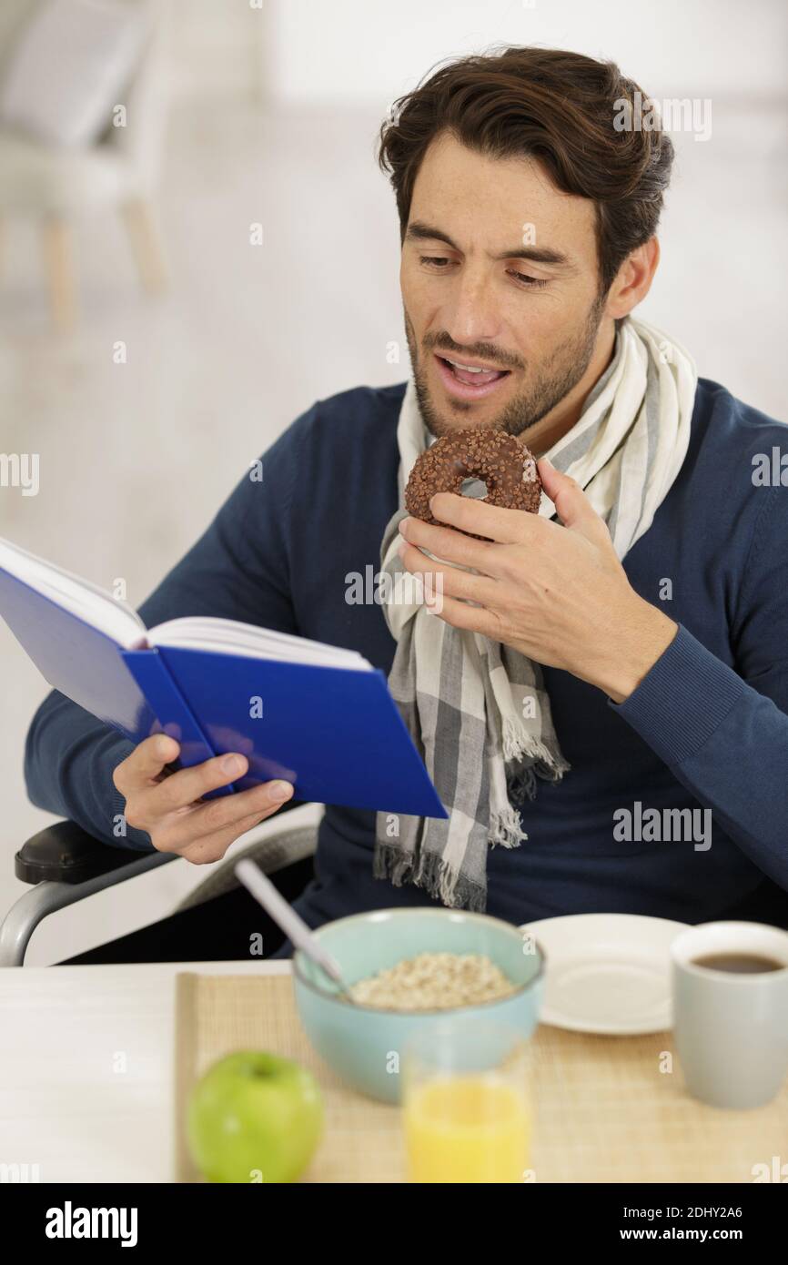 man in wheelchair reading book and eating chocolate donut Stock Photo