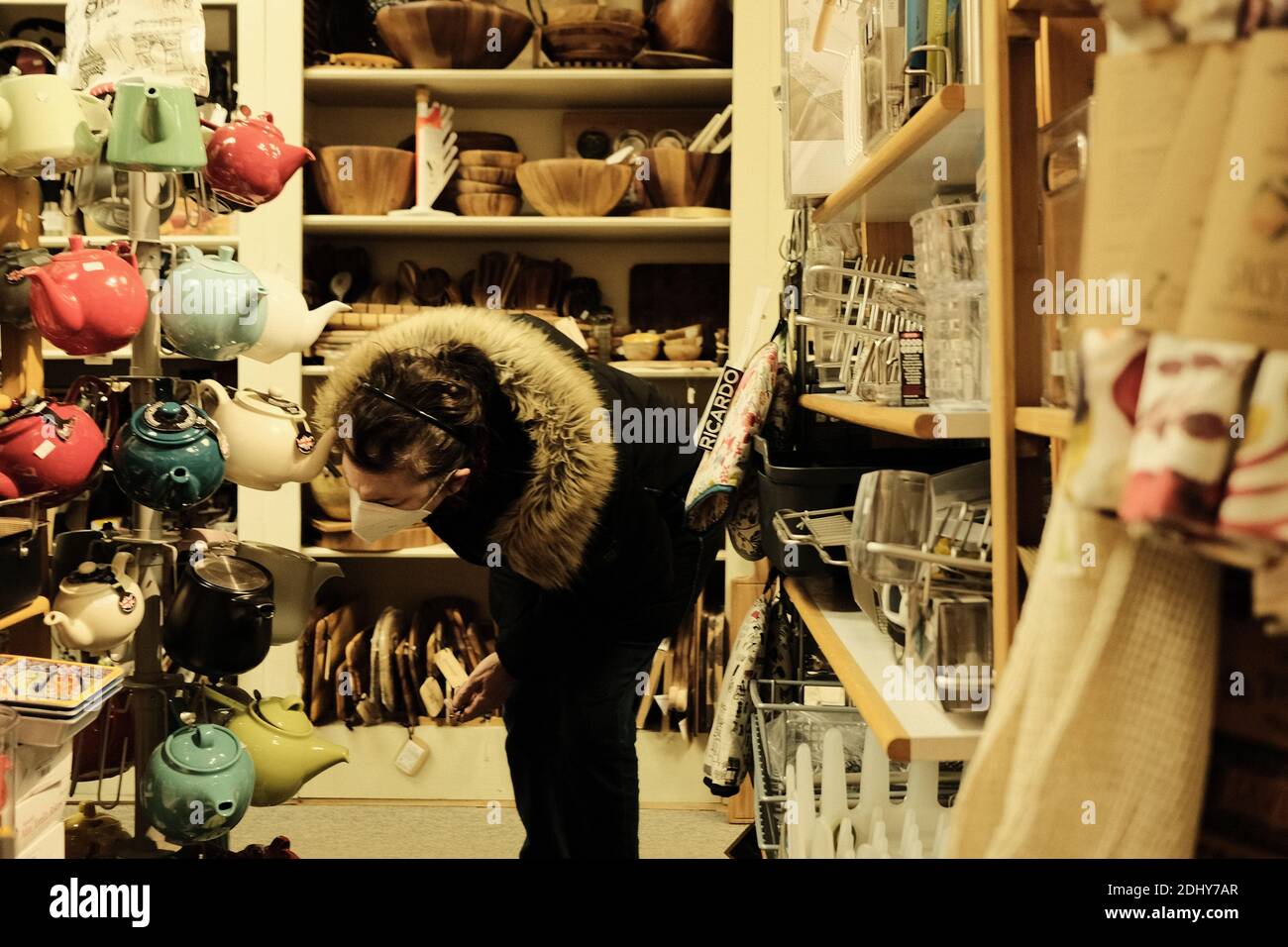Masked shopper in a winter jacket examines teapots in s small kitchen