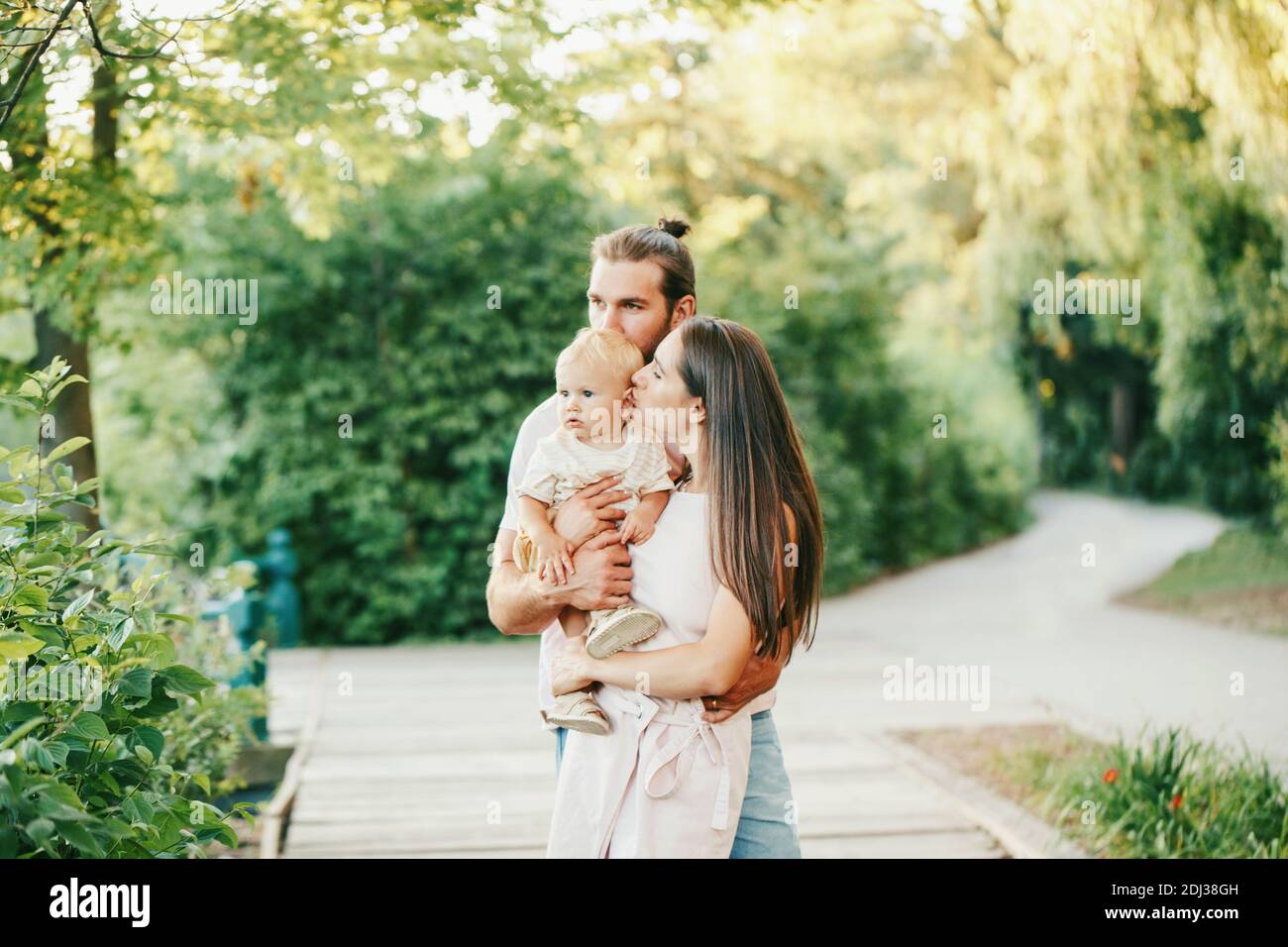 Family Day. Mother and father with baby boy in park outdoor. Family Caucasian mom and dad with son walking hugging in park on spring summer day. Happy Stock Photo