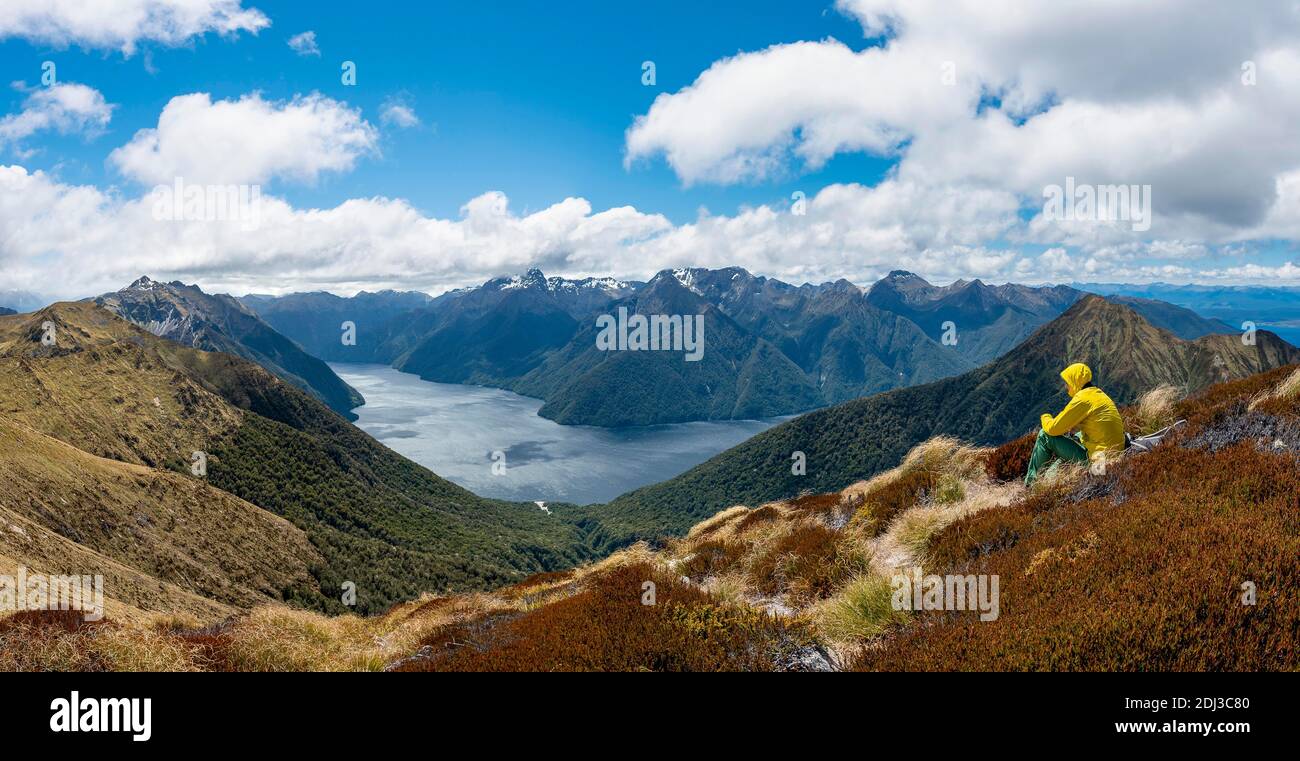 Mountaineer, hiker sitting in the grass, view of the South Fiord of Lake Te Anau, Murchison Mountains and Southern Alps in the background, on the Stock Photo