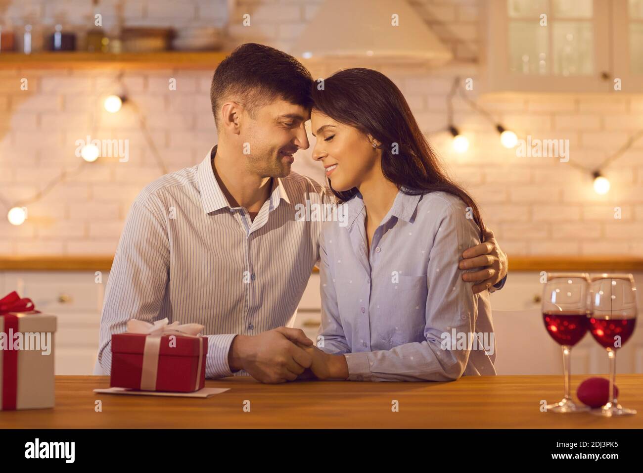 Young loving couple sitting at table with wine and holiday presents, hugging and celebrating Valentines day Stock Photo