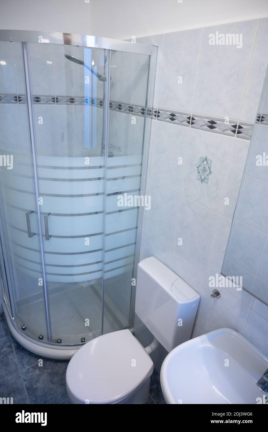Interior of a narrow and long bathroom, the glass shower enclosure with metal frame is an eye-catcher. White ceramic sanitary ware. Stock Photo