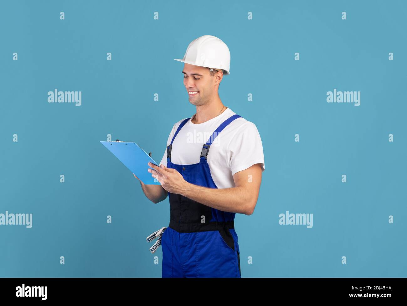 Smiling male repair contractor in coveralls and hardhat writing notes in clipboard Stock Photo
