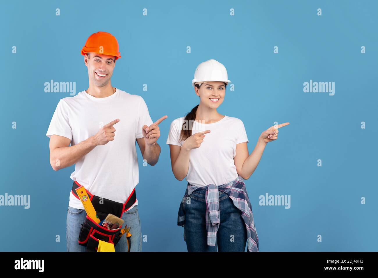 Smiling handyworkers couple wearing casual clothes and protective hard hats pointing aside Stock Photo